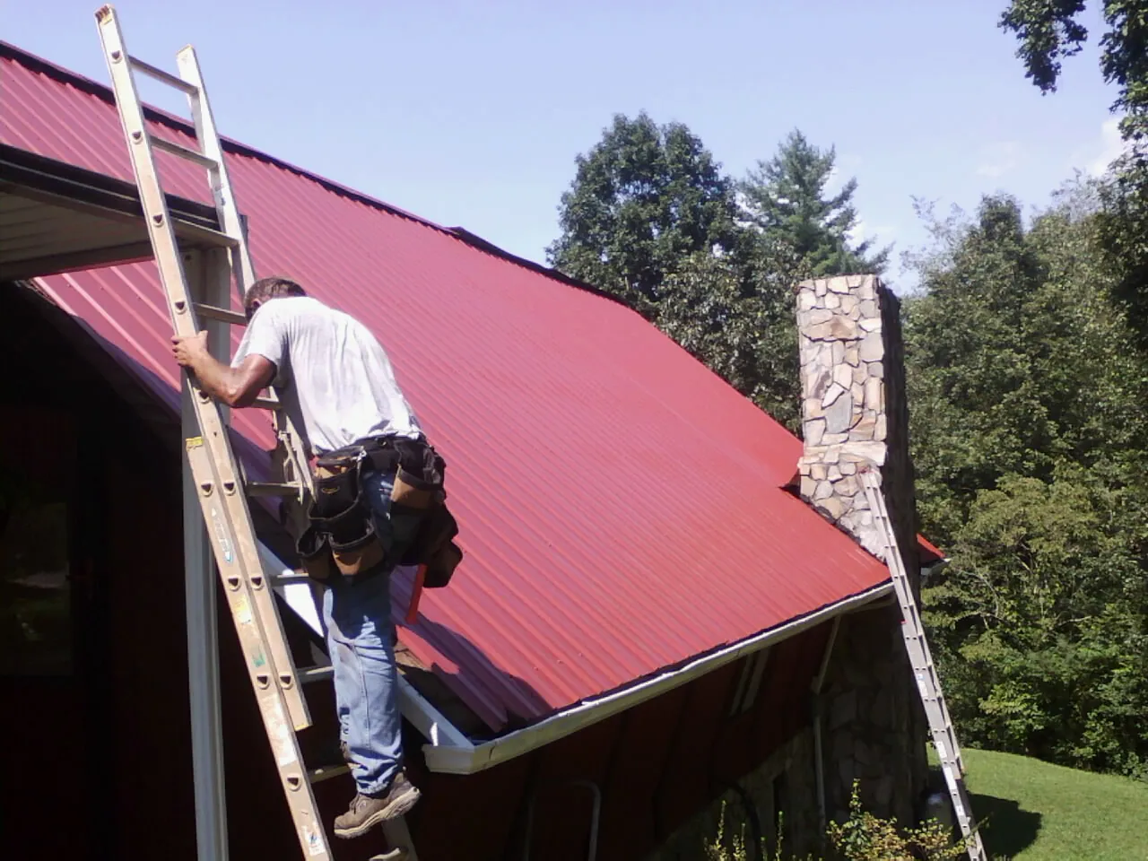 Roofer on a ladder beside a red metal roof and stone chimney — Soffit & Fascia Repair by Roofing Brothers in Redmond